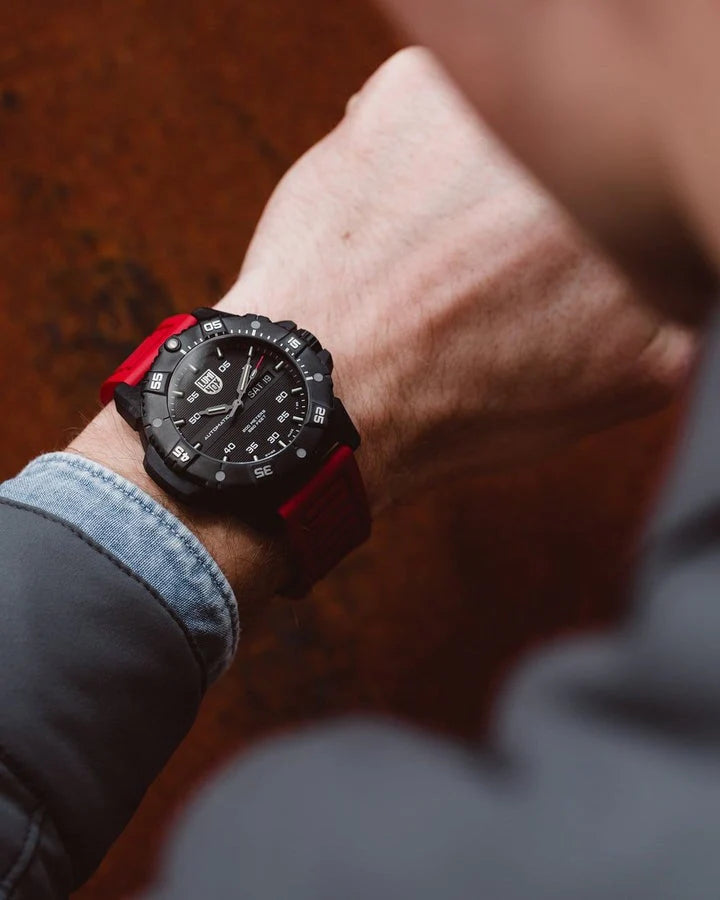 Black wristwatch with a red strap worn on a person’s wrist.
