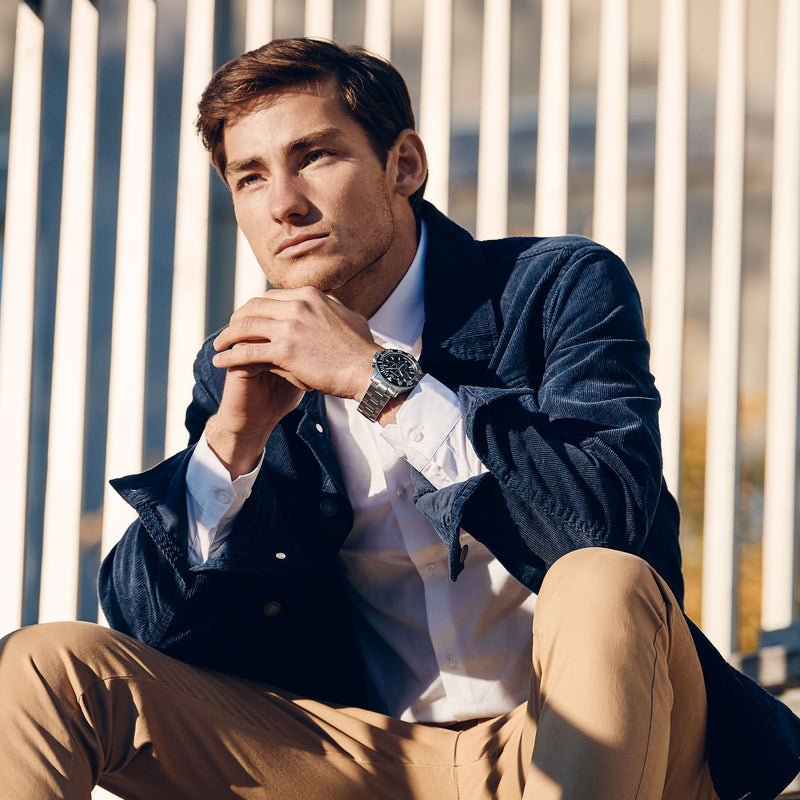 Man wearing a navy jacket and khaki pants, sitting with a thoughtful expression.