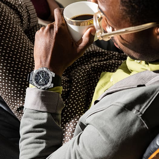 Wristwatch with a black face and silver metal band worn by someone drinking coffee.