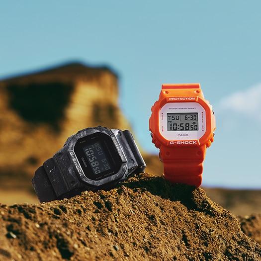 Two G-Shock digital watches, one black and one orange, sitting on a rocky surface.