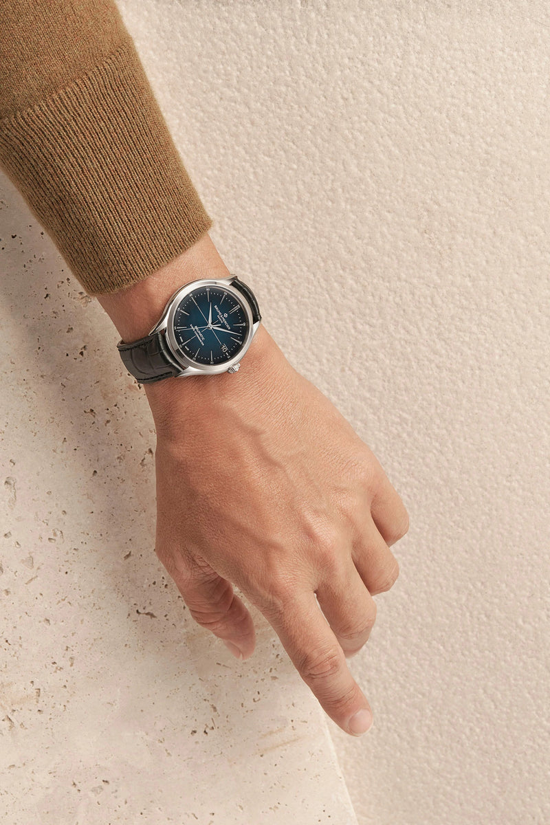 Wristwatch with a dark face and gray leather strap worn on a person’s wrist.